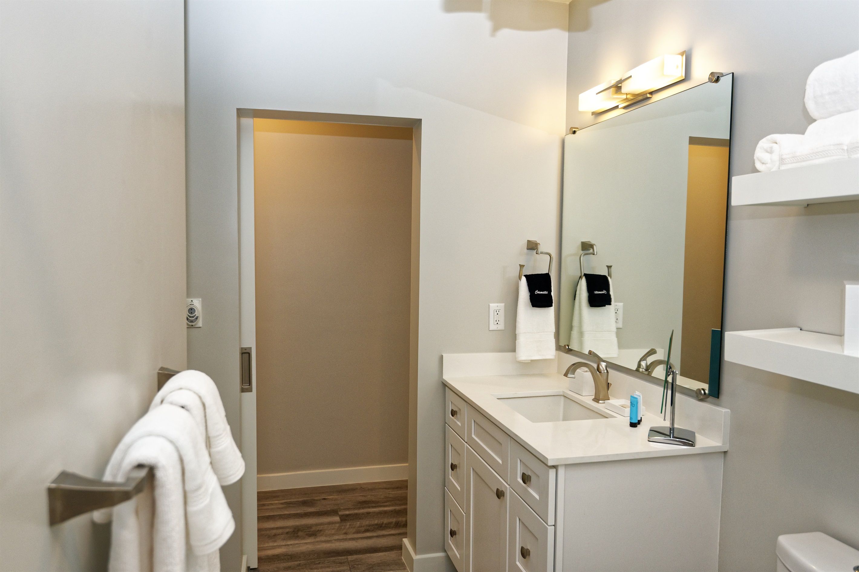 A modern bathroom with a large mirror, a sink, and a toilet. There is a towel rack with a white towel and a black towel with the word 'Cosmetic' on it.