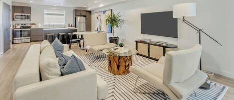 Una sala de estar moderna con un sofá, una mesa de centro y un televisor. Hay una zona de cocina con un microondas y una mesa de comedor.