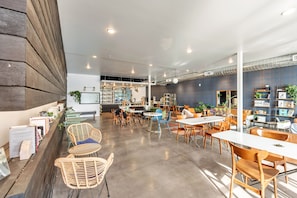 Un café spacieux doté de plusieurs espaces de détente, comprenant des tables et des chaises, ainsi qu’un bar avec des étagères remplies de livres et de plantes.