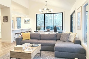 A modern living room with a grey sectional sofa, a wooden coffee table, and a large window with a view of trees. - Walk to the Lifts! Park City 3BR Condo w/ Garage (Park City)