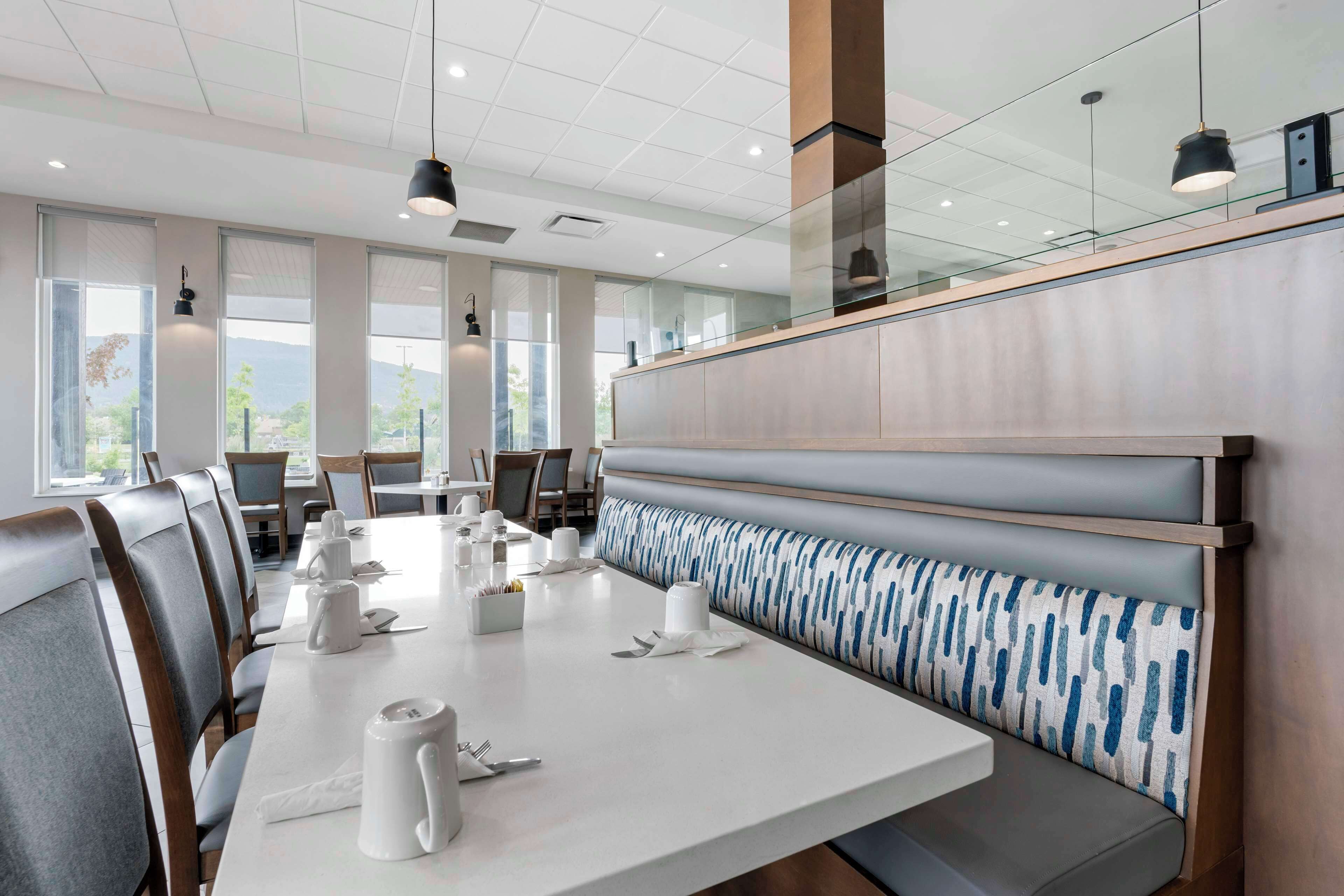 A modern dining area with a long table set for a meal, cushioned seating, and large windows offering a view.