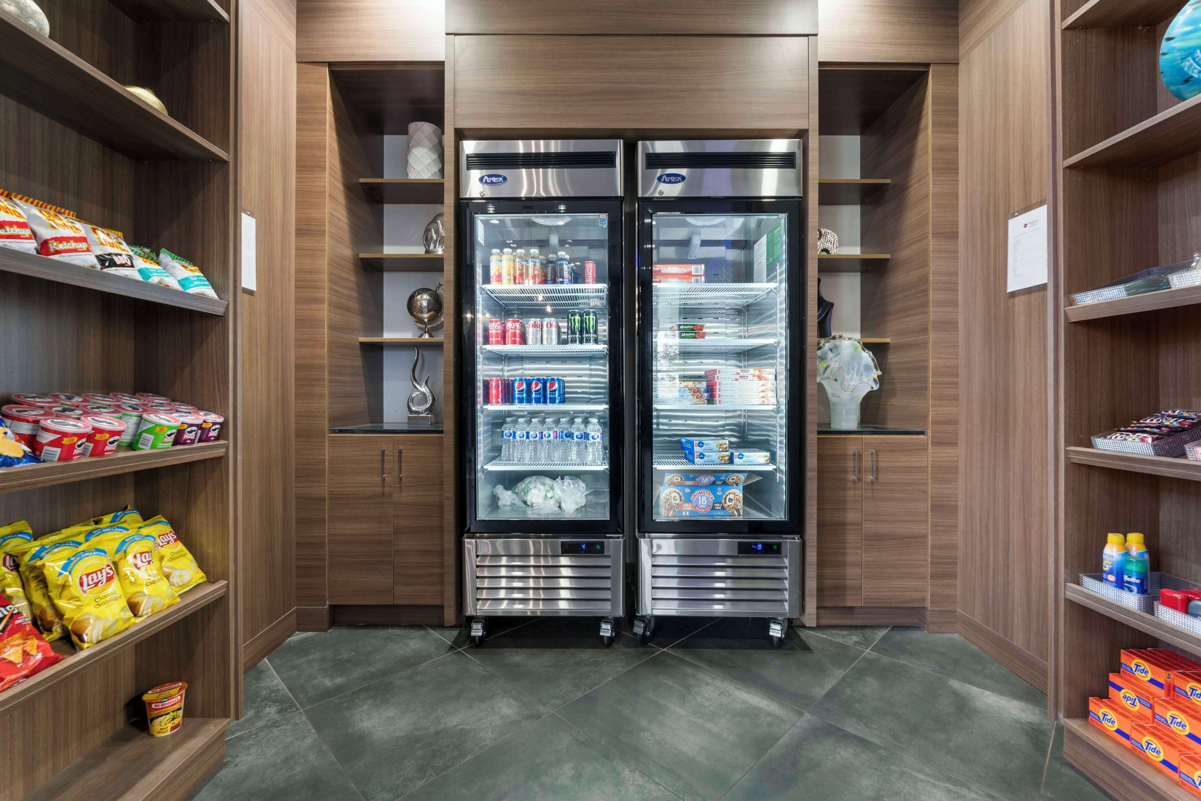 Una kitchenette moderna y compacta con dos refrigeradores de acero inoxidable, provista de bebidas y aperitivos, y muebles de madera con estantes que contienen diversos alimentos.