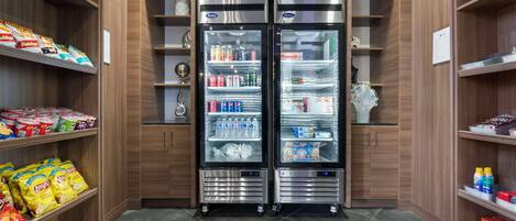 Una kitchenette moderna y compacta con dos refrigeradores de acero inoxidable, provista de bebidas y aperitivos, y muebles de madera con estantes que contienen diversos alimentos.