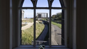 Une vue à travers une fenêtre ornée d’une arche gothique, donnant sur un chemin pavé et un bâtiment doté de colonnes.