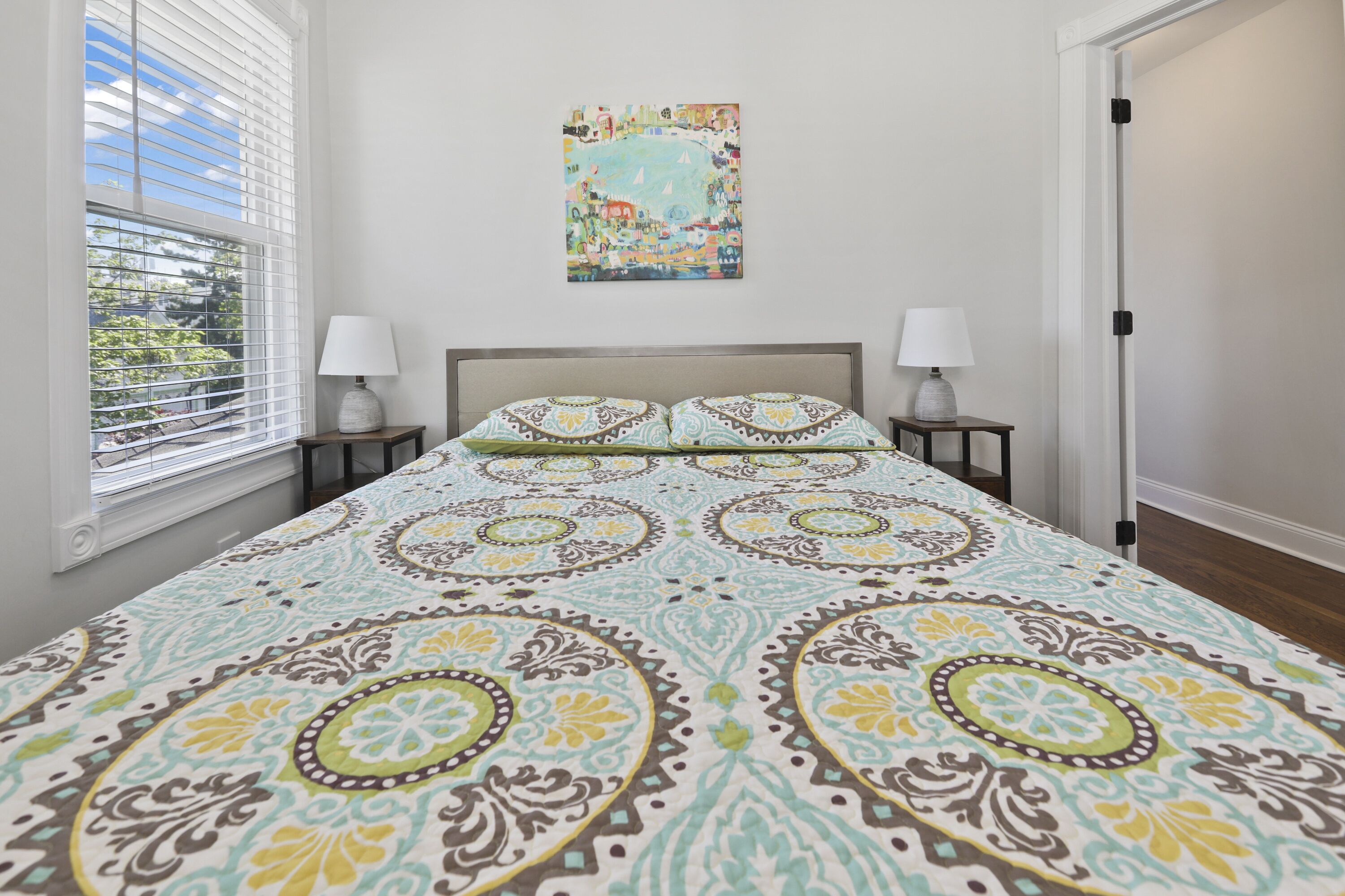 A bedroom with a large bed, two bedside tables, a window with blinds, and a colorful wall hanging.