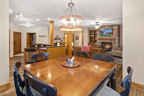 Un comedor con una mesa y sillas de madera, una araña, una cocina al fondo y una chimenea de piedra con un televisor encima.