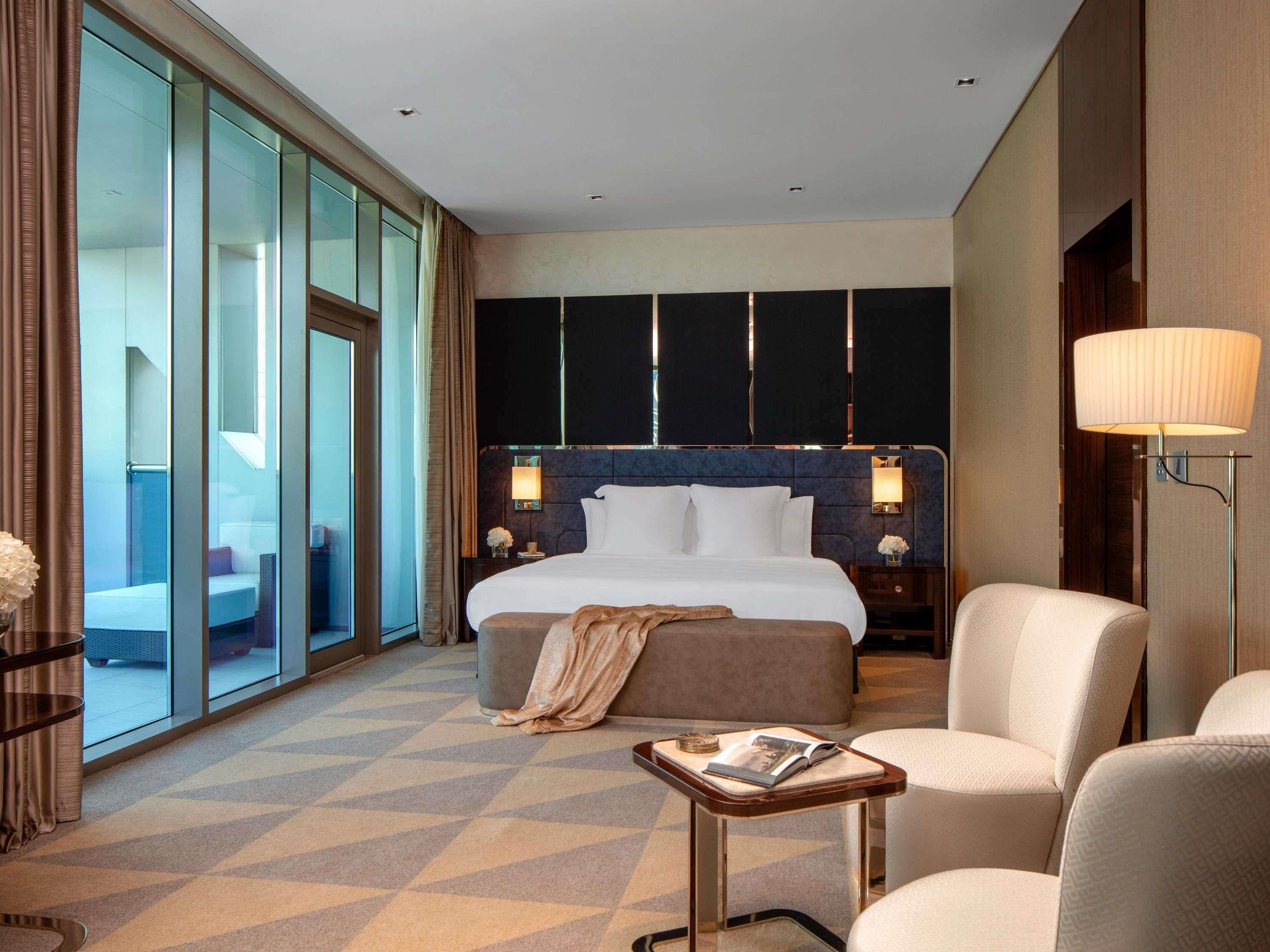 A hotel room with a large bed, a seating area with a table, and a view through floor-to-ceiling windows.