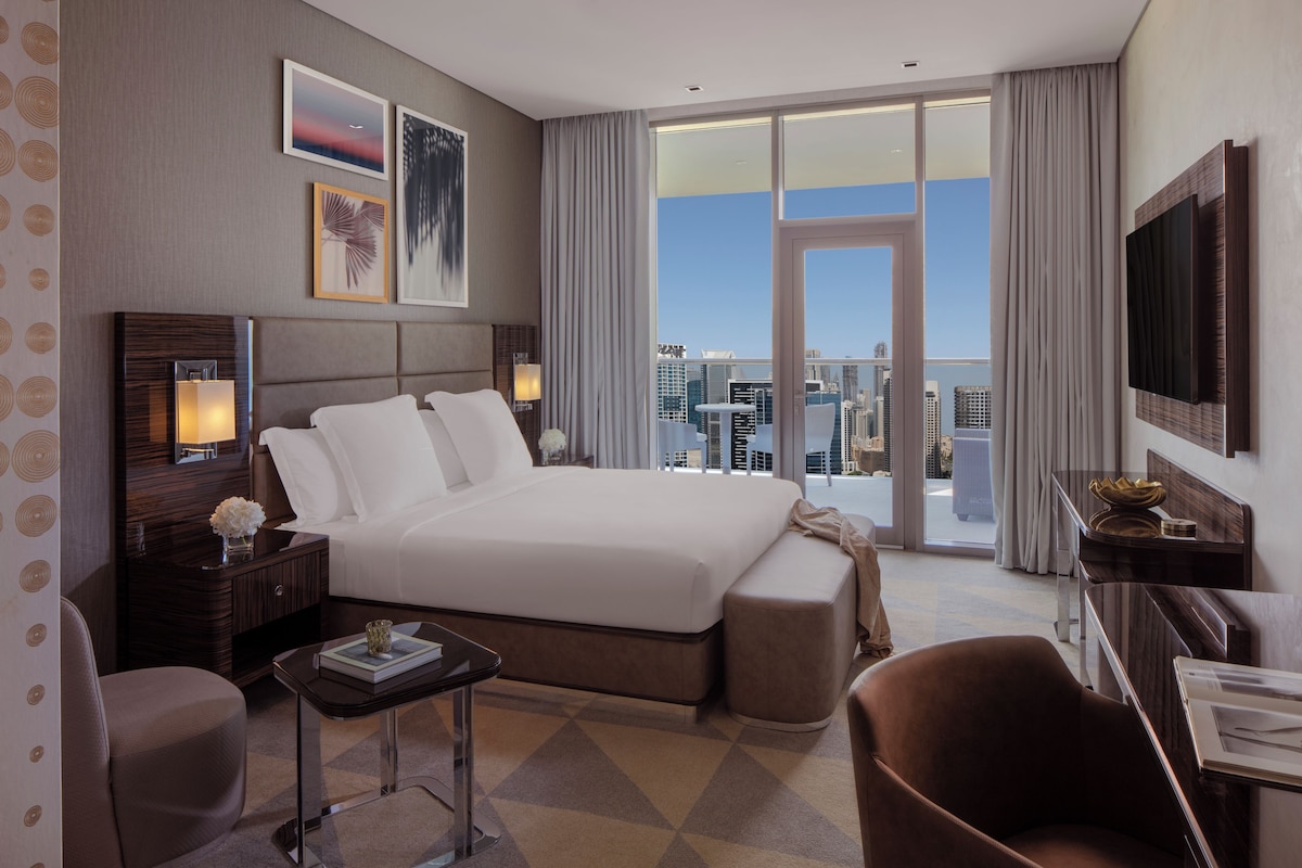 A hotel room with a large bed, a desk, a chair, a TV, and a view of the cityscape.