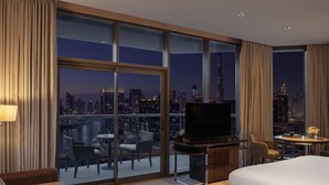 A hotel room with a large window, a bed, a TV, a desk, and a balcony with a city view.