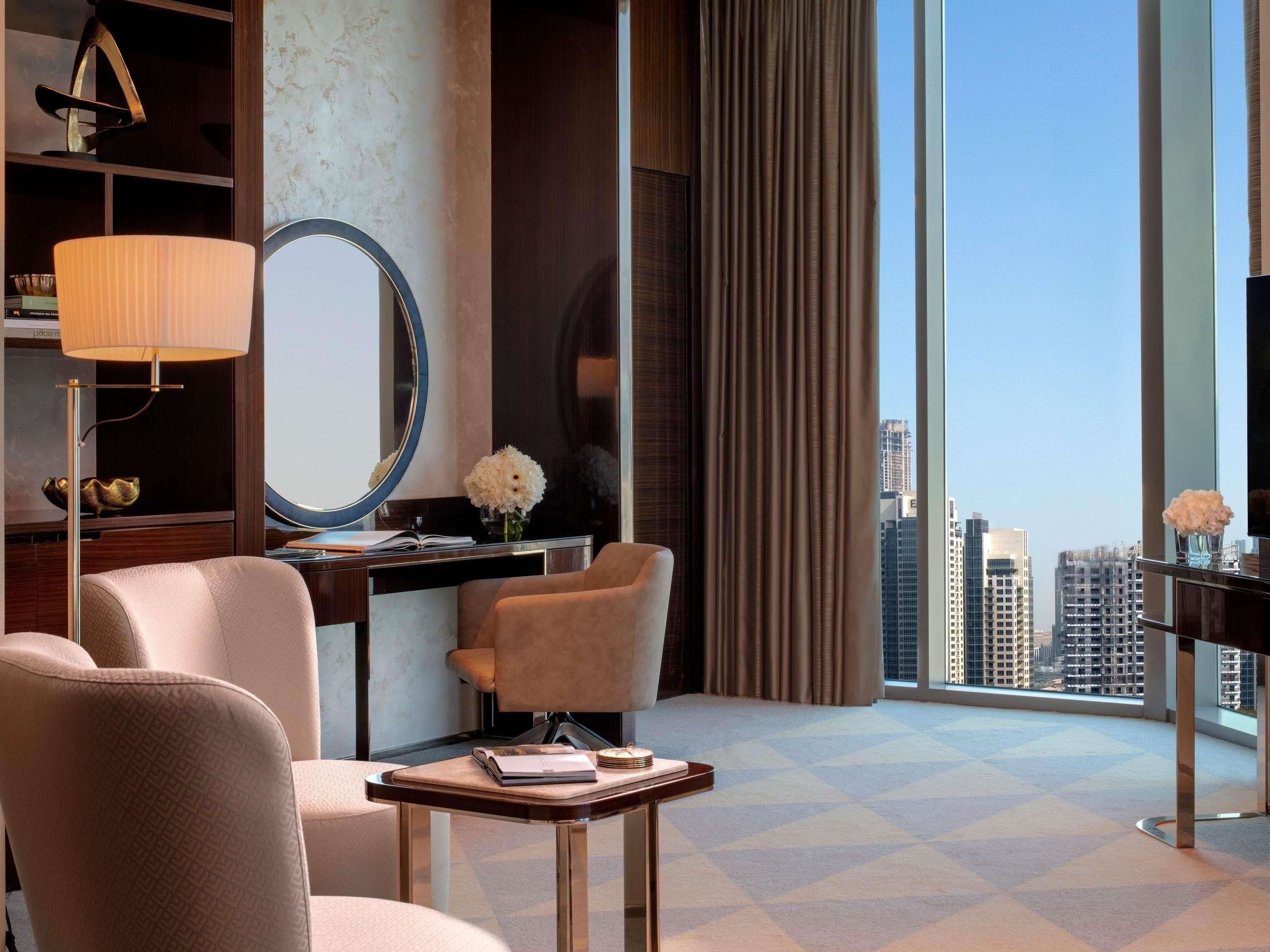 A hotel room with a city view, a desk with a mirror, a lamp, and two chairs.