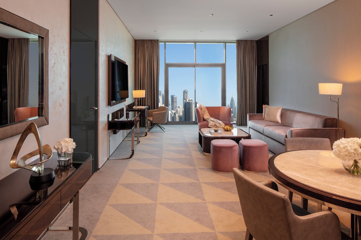A hotel room with a city view, a flat-screen TV, a desk with a lamp, a sofa, a round dining table, and a chair.
