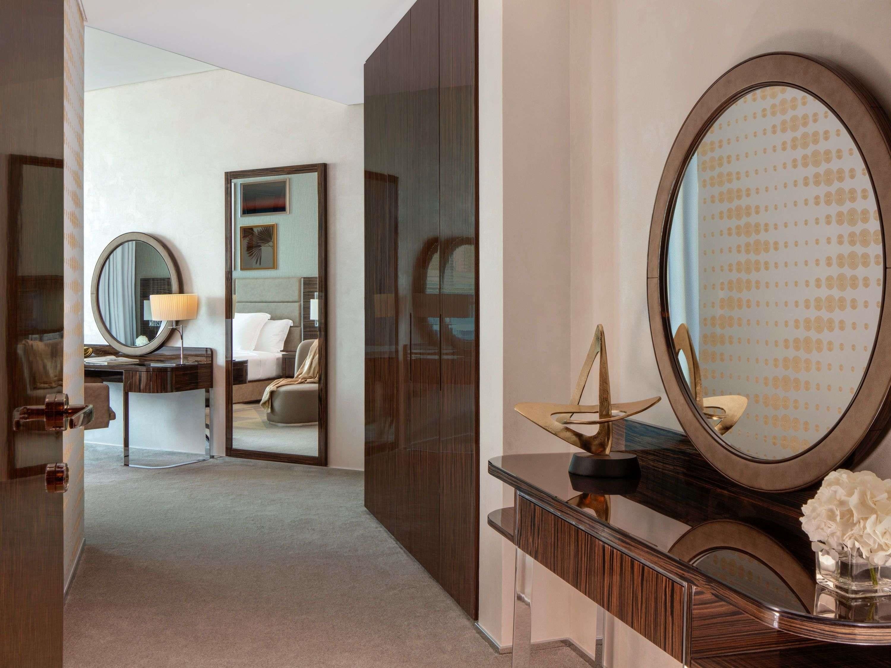 A modern hotel room with a large mirror, a desk with a lamp, a comfortable bed, and a decorative vase.