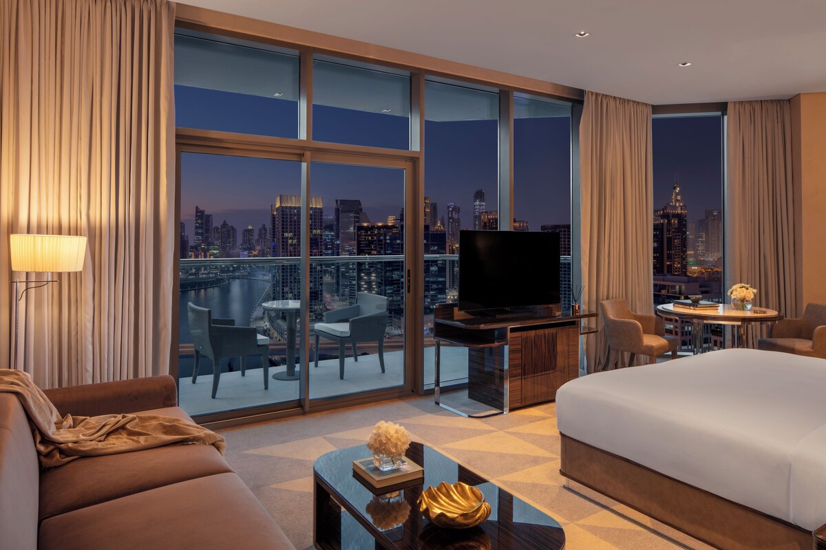 A hotel room with a large bed, a TV, a seating area with a table and chairs, and a view of a cityscape at night.