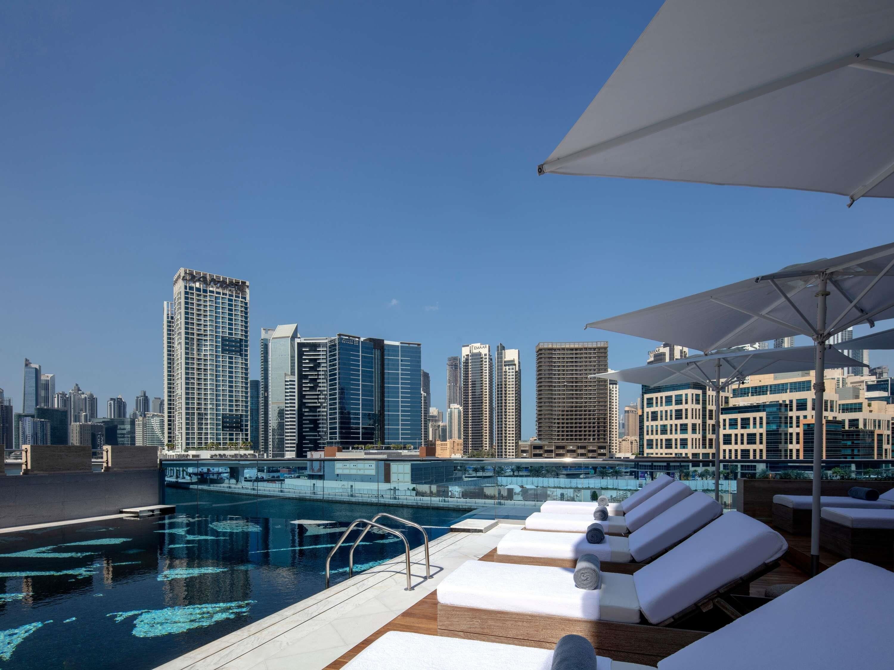 Un área de piscina en la azotea con sillones de descanso y sombrillas, con vistas a un horizonte urbano.