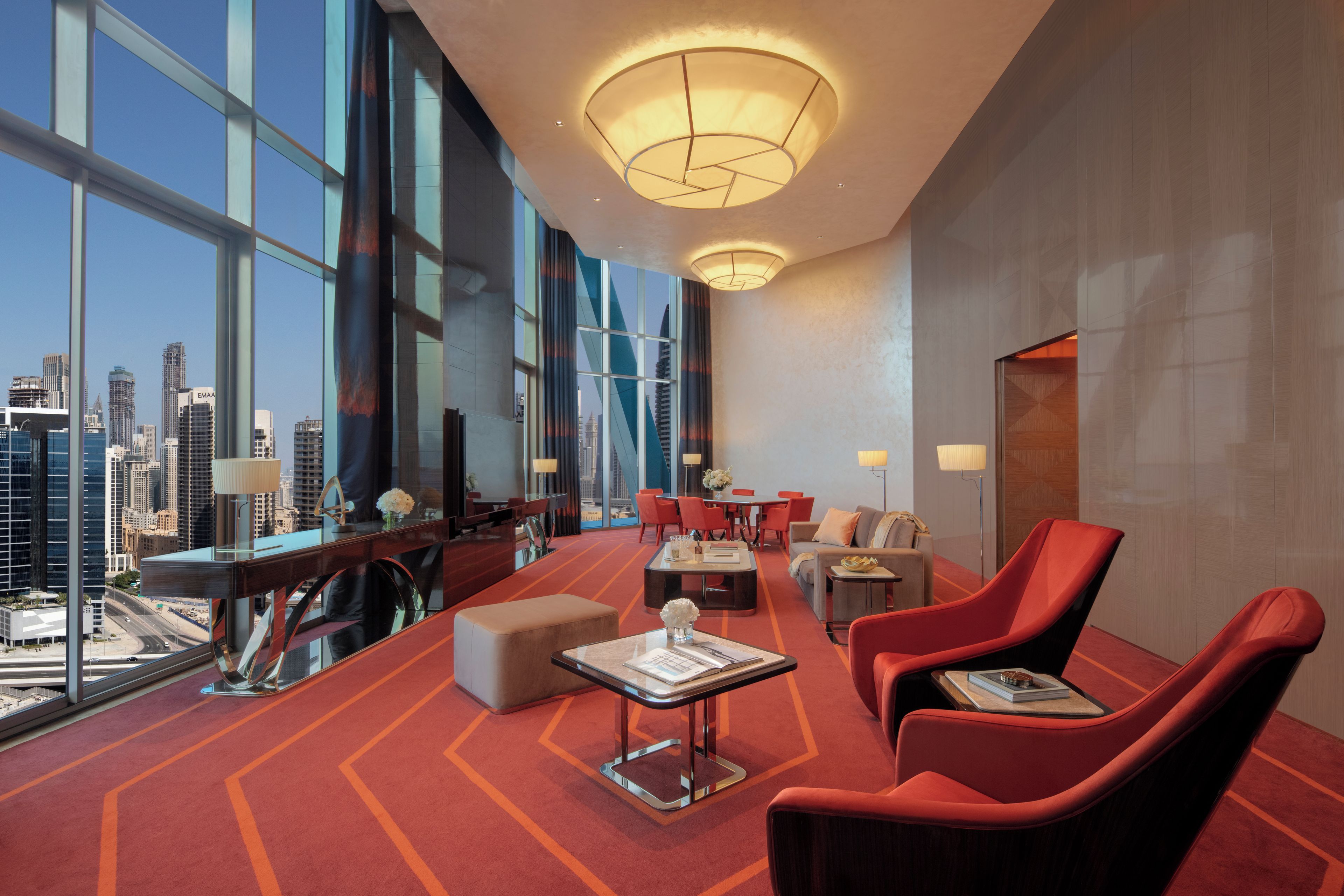 Una moderna hall d'albergo con vista panoramica sulla città, un'area salotto centrale con poltrone rosse e un grande lampadario.