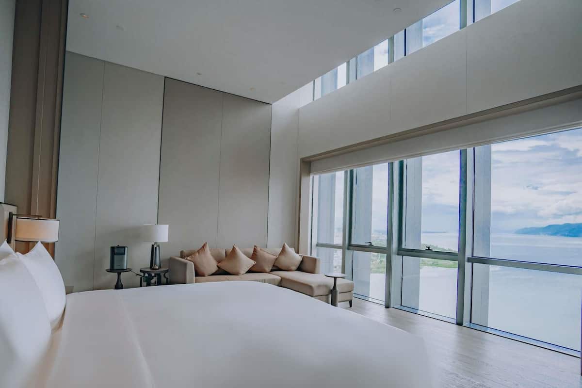 A modern hotel room with a large bed, a sofa, a desk, and a view of the sea.
