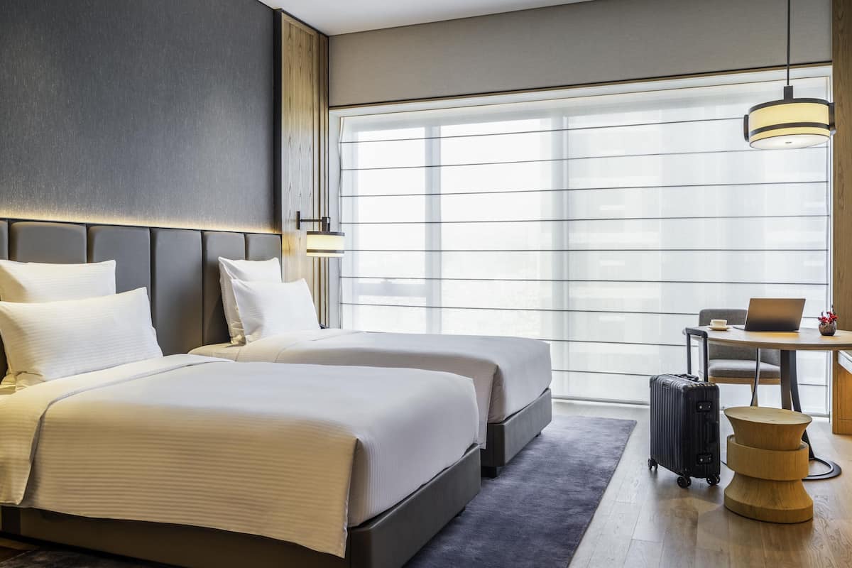 A hotel room with two beds, a desk with a laptop, a suitcase, and a large window.