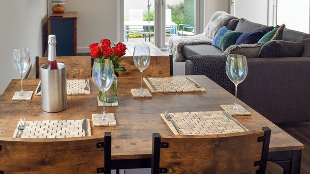 A dining area with a wooden table set for four, including wine glasses, cutlery, and a bottle of red wine. A vase with red roses is on the table. - 1 bedroom accommodation in St Andrews (St Andrews)