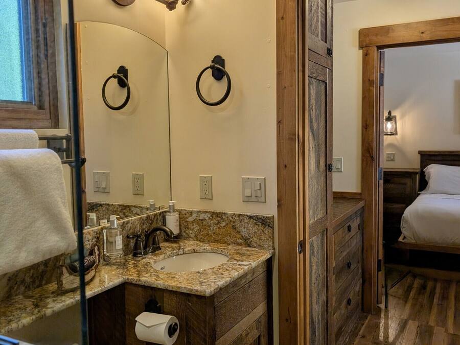 Ein Badezimmer mit Granitplatte, Holzschränken, einem beleuchteten Spiegel, einem Handtuchhalter und einem Bett im angrenzenden Raum.