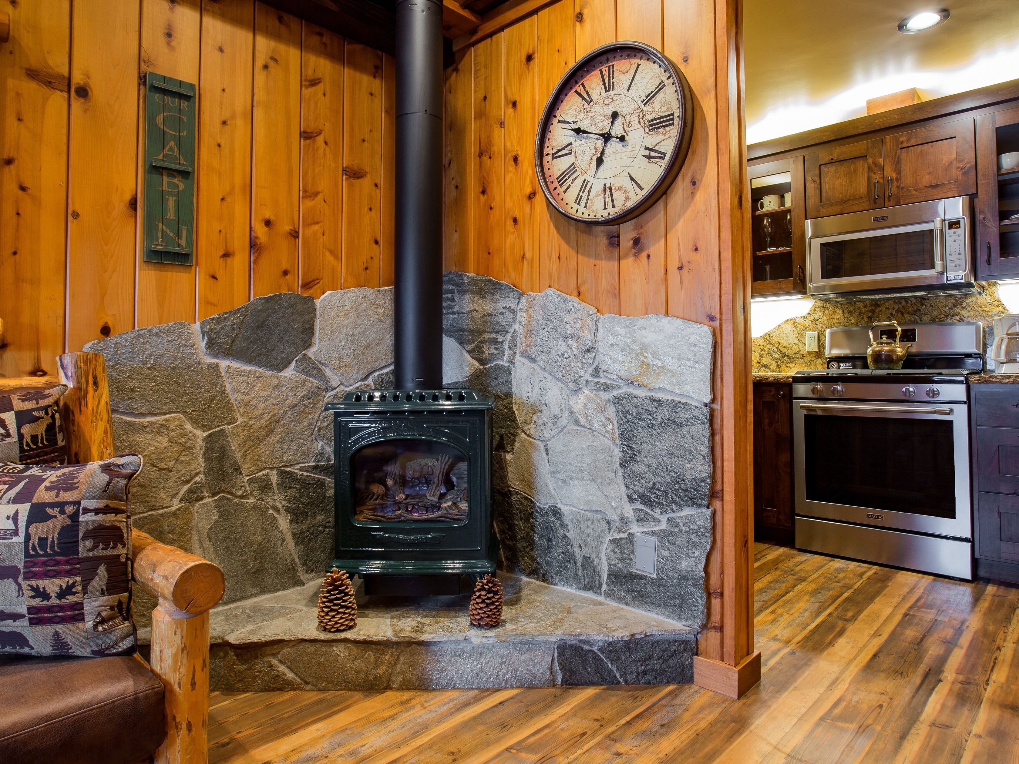 Ein gemütliches Blockhäuschen-Interieur mit Kamin aus Naturstein, Holzwänden, einer Uhr und modernen Küchengeräten.
