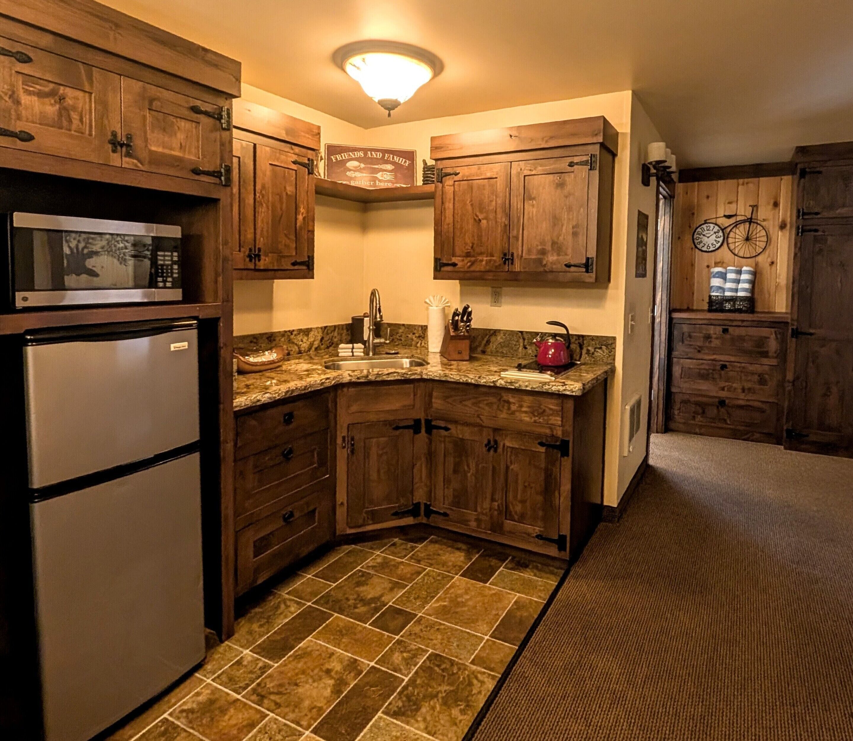 Una cocina con armarios de madera, microondas, refrigerador, lavamanos y un reloj en la pared.