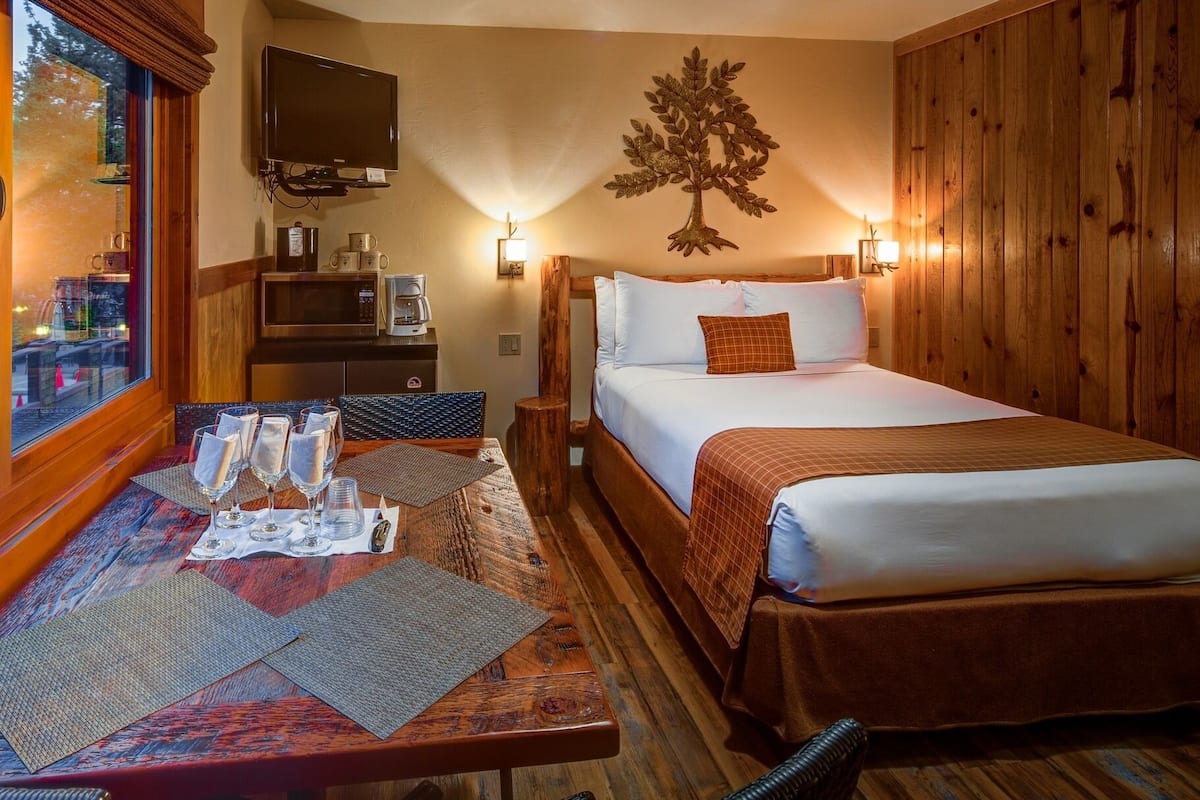 Ein Hotelzimmer mit Bett, Schreibtisch mit Gläsern und einer Kaffeemaschine, Fernseher und einer Holzwand mit Baumdeko.