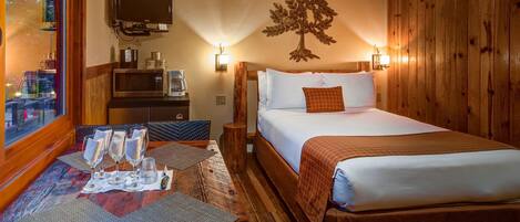 Una habitación de hotel con cama, escritorio con vasos y cafetera, televisor y pared de madera decorada con un árbol.