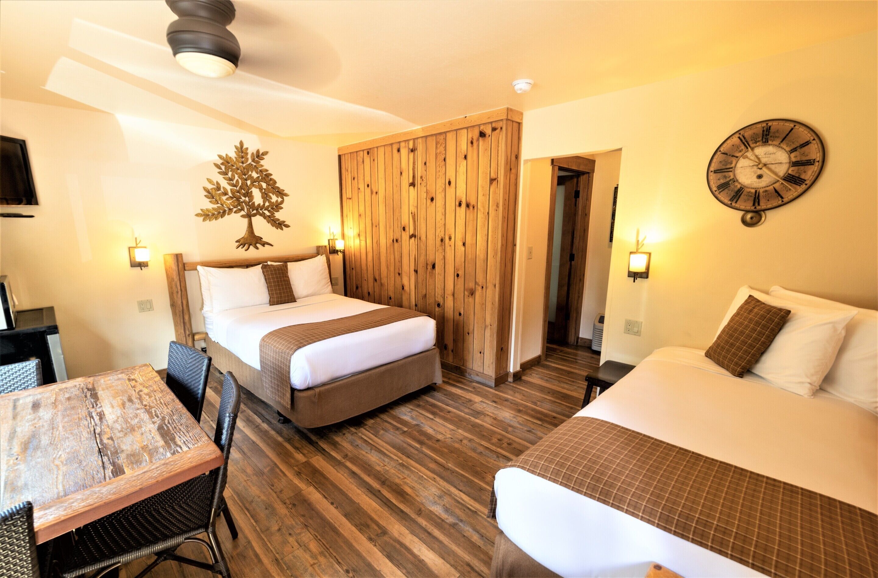 A hotel room with two beds, a wooden wardrobe, a dining table with chairs, and a wall clock.
