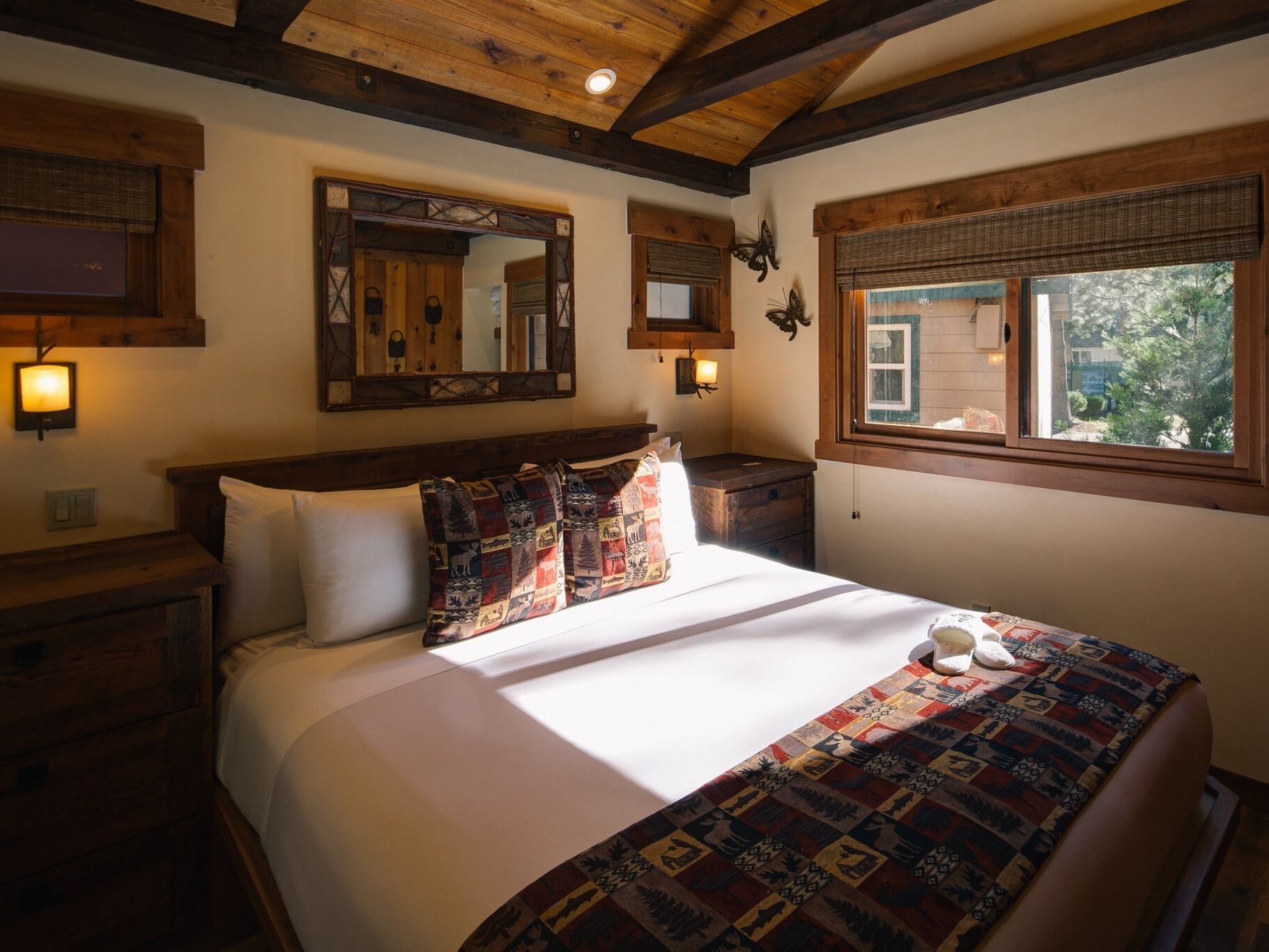 A bedroom with a wooden ceiling, a large bed with patterned bedding, a nightstand, a mirror, and a window with a view of greenery.