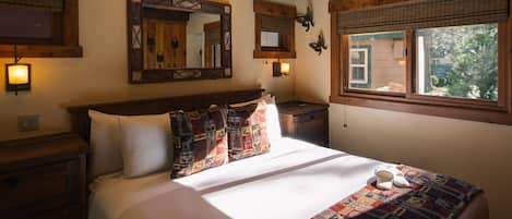 Un dormitorio con techo de madera, una cama grande con ropa de cama estampada, una mesita de noche, un espejo y una ventana con vistas a la vegetación.