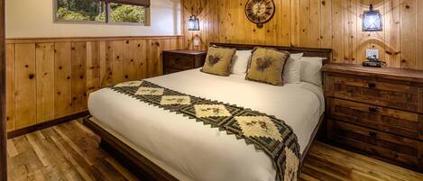 A bedroom with a wooden paneled wall, a large bed with a quilt, two bedside tables with lamps, and a window with blinds.