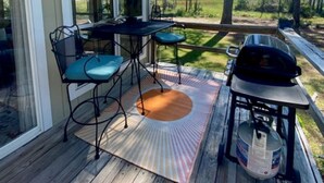 A wooden deck with a table and chairs, a grill, and a large window overlooking a grassy area. - Marsh Beauty (Alligator Point)