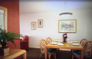 Une salle à manger avec une table en bois dressée pour quatre personnes, entourée de chaises en osier. Des tableaux encadrés ornent le mur au-dessus de la table.