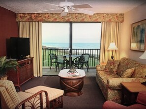 Une pièce comprenant une télévision, un canapé, un fauteuil, une petite table et un balcon avec vue sur l’océan.