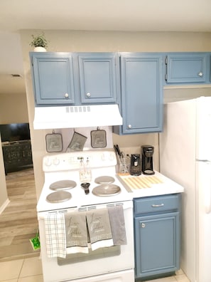 Una cocina con armarios azules, una estufa blanca y un refrigerador. Hay utensilios de cocina y una cafetera en la encimera.