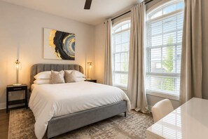 A modern bedroom with a large bed, bedside lamps, a painting, and a view through large windows. - Cozy Nashville Home  • Walk to Vandy (nashville)