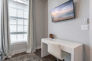Una habitación con un ventanal grande, un televisor de pantalla plana montado en la pared, un escritorio blanco y un pequeño adorno sobre el escritorio.
