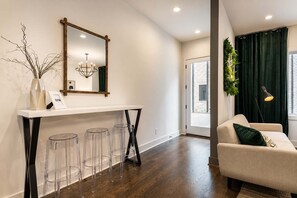 A modern living room with a sofa, a coffee table, and a lamp. There is a dining area with a white console table and clear stools. A large mirror hangs above the console table.