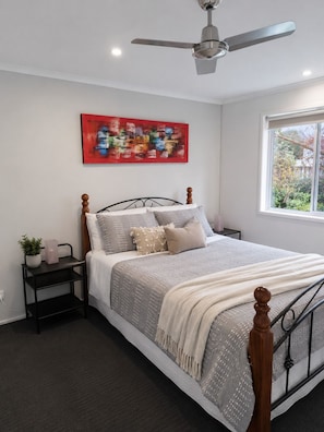 Ein Schlafzimmer mit Bett, Deckenventilator, Fenster, Nachttisch mit Pflanze und einem Gemälde an der Wand.