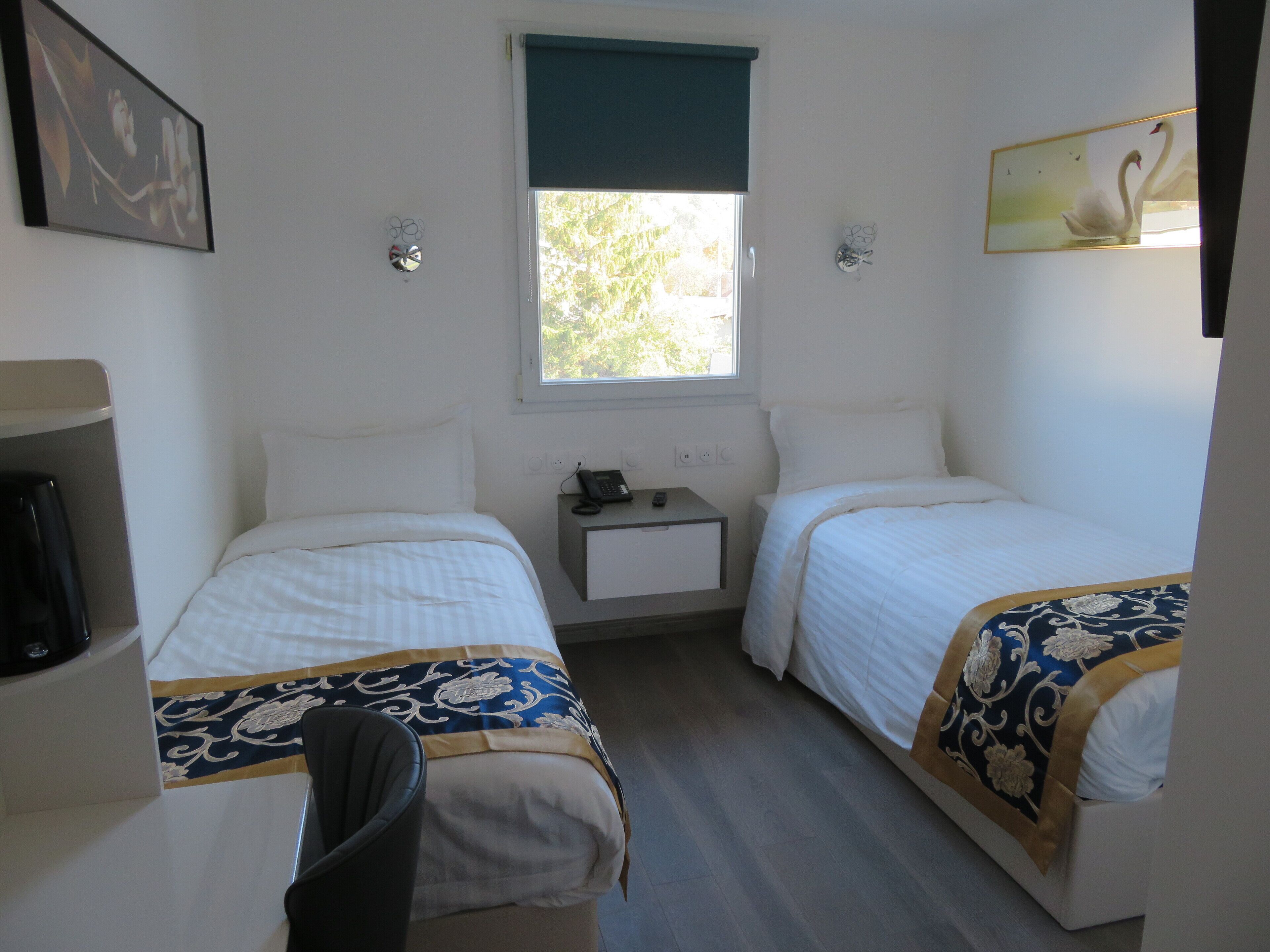 Una habitación pequeña y limpia con dos camas, un escritorio, una silla, una ventana con vistas a árboles y un cuadro de un cisne.