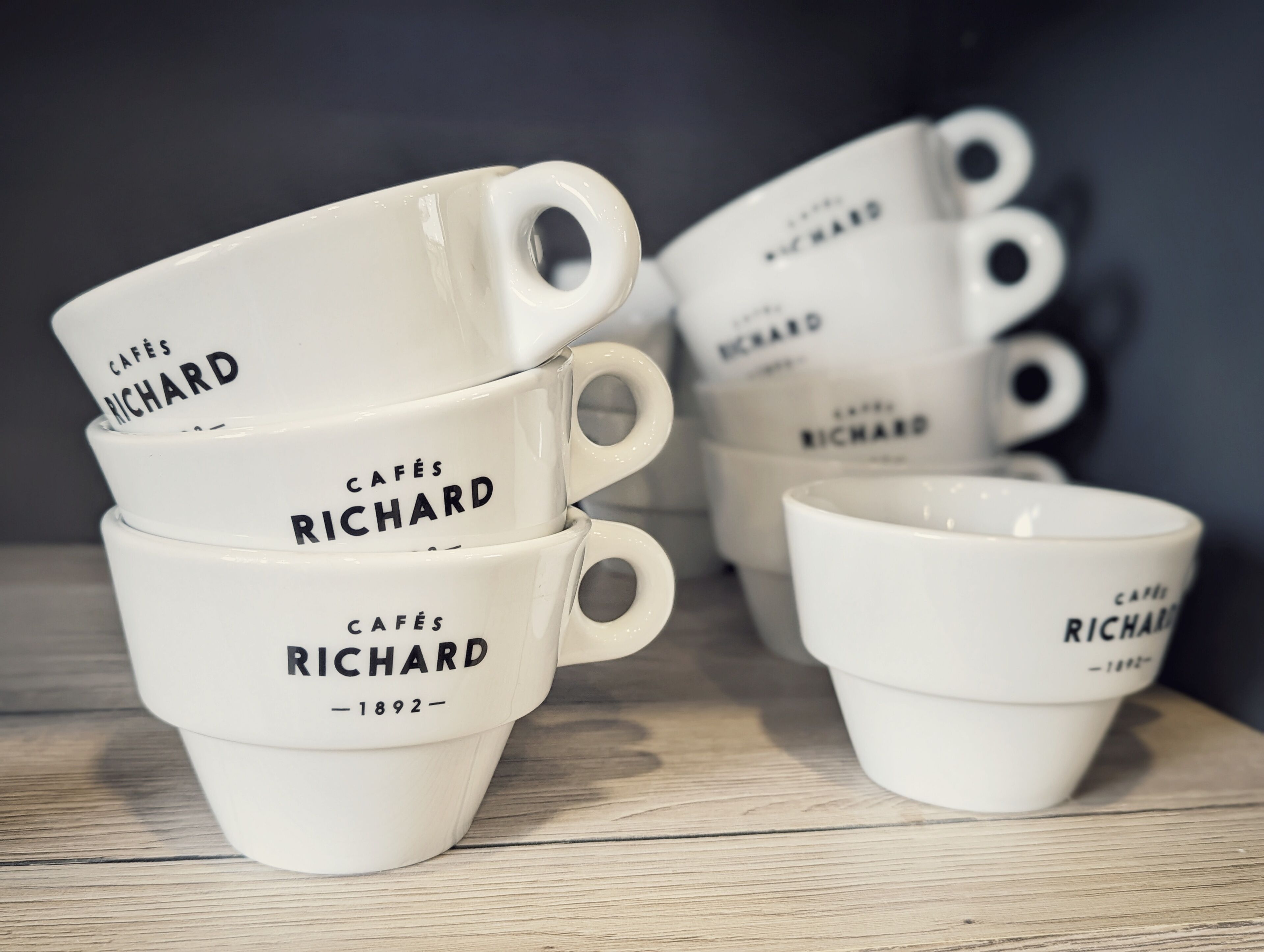 A stack of white coffee cups with the text "Cafés Richard - 1892" on them, placed on a wooden surface.