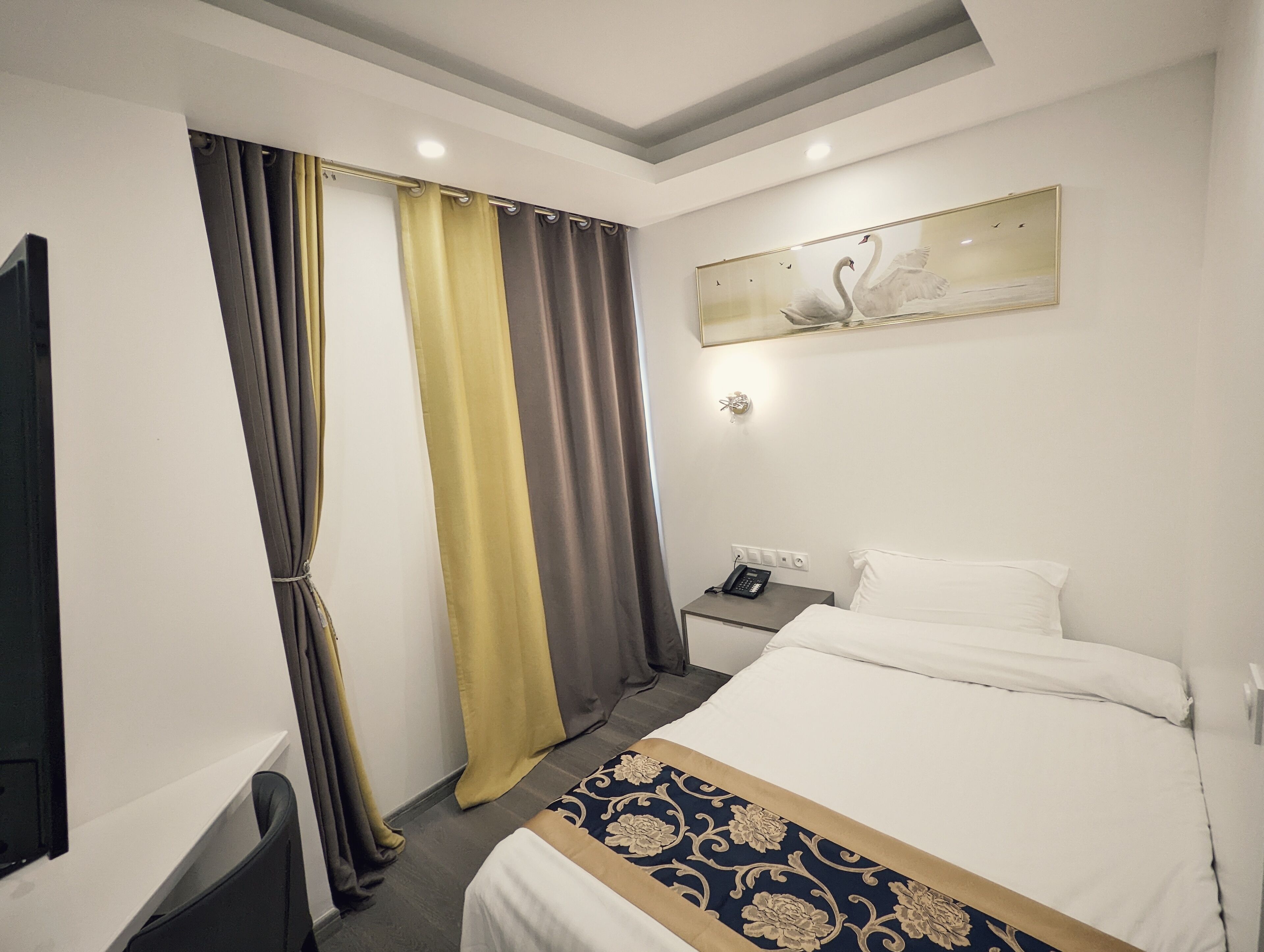 A hotel room with a bed, a bedside table with a phone, a wall-mounted light, and a painting of swans.