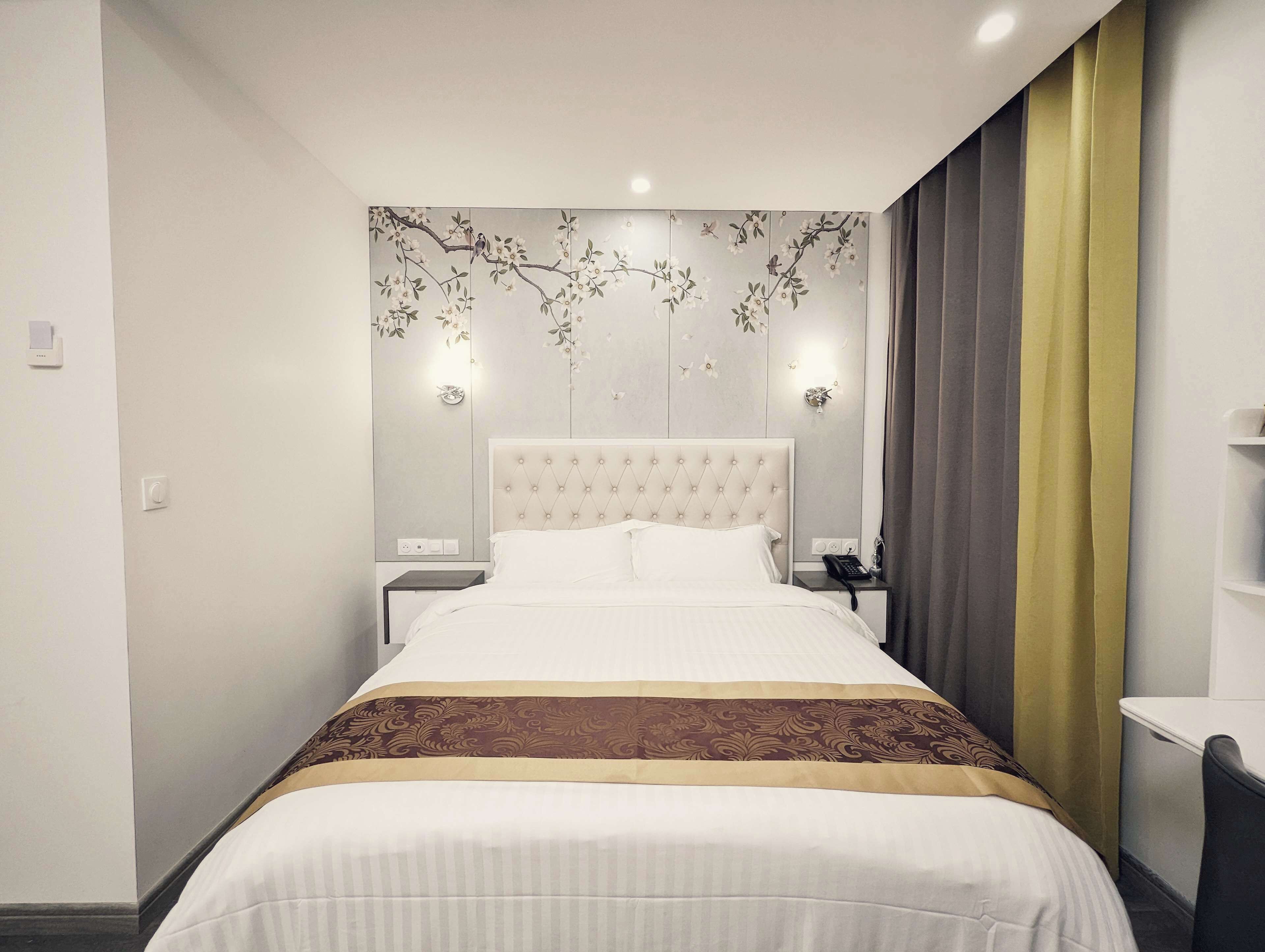 A modern hotel room with a large bed, a nightstand, a bedside table, and a wall-mounted light fixture.