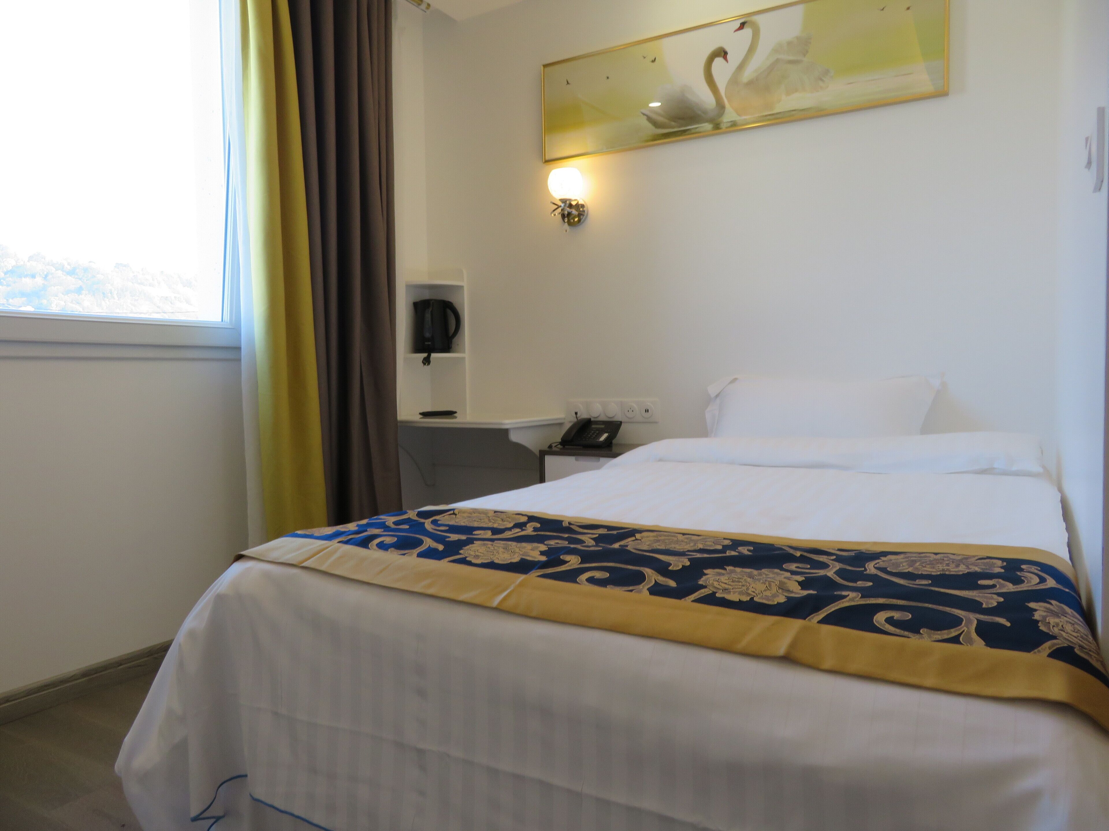 A hotel room with a bed, a bedside table with a phone, a wall-mounted light, and a window with curtains.