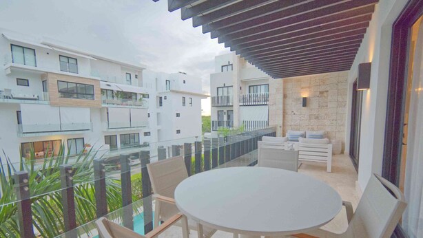 Un patio exterior moderno con una mesa redonda y sillas, rodeado de barandillas de vidrio, con vistas a una piscina y edificios cercanos.