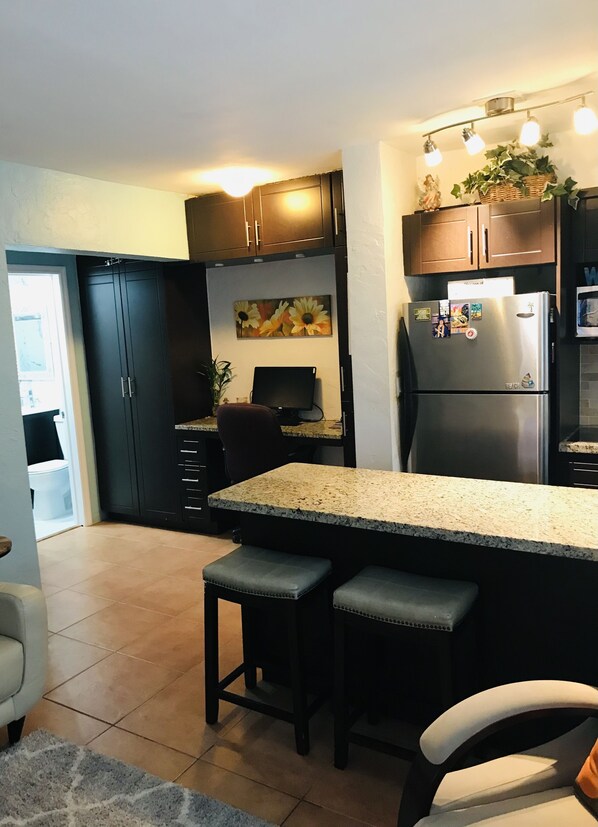 A modern kitchen with dark cabinets, a granite countertop, two bar stools, a refrigerator, and a dining table.