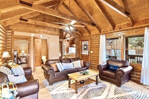 L’intérieur d’une cabane en rondins, avec un plafond en bois, un ventilateur de plafond, un canapé en cuir marron, une table basse en bois et une vue par la fenêtre.