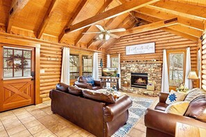A log cabin interior with a fireplace, leather sofas, wooden beams, and a stone wall. - 2Mi to Dollywood • Arcade Games • 3BR • SmartTVs • HotTub • BBQ • Easy Access!! (Pigeon Forge)