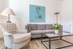 A modern living room with a grey sofa, a tufted armchair, a glass coffee table, a floor lamp, and a wall art piece. - Stimmig. Besonders. Exklusiv. - Art Your Life - Residenz, Whg. 6 "craig" - Objekt-id: Rsw/06 - Art (Sylt)