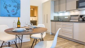Una cocina moderna con armarios de colores claros, un microondas integrado y una cafetera. Hay un comedor con una mesa redonda y sillas blancas. Un cuadro con un diseño abstracto azul está en la pared.